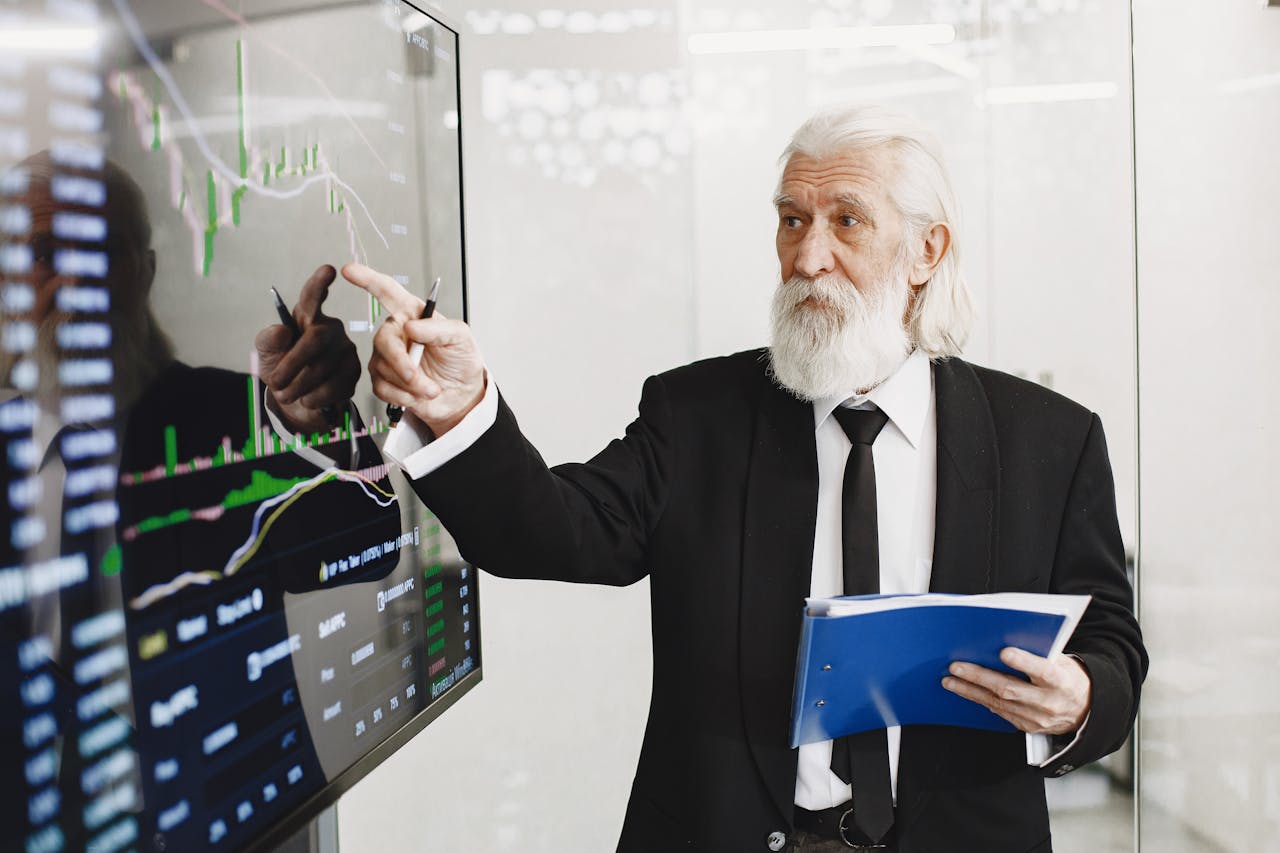 services-03 Elderly businessman pointing at stock market charts in an office setting, highlighting data analysis.